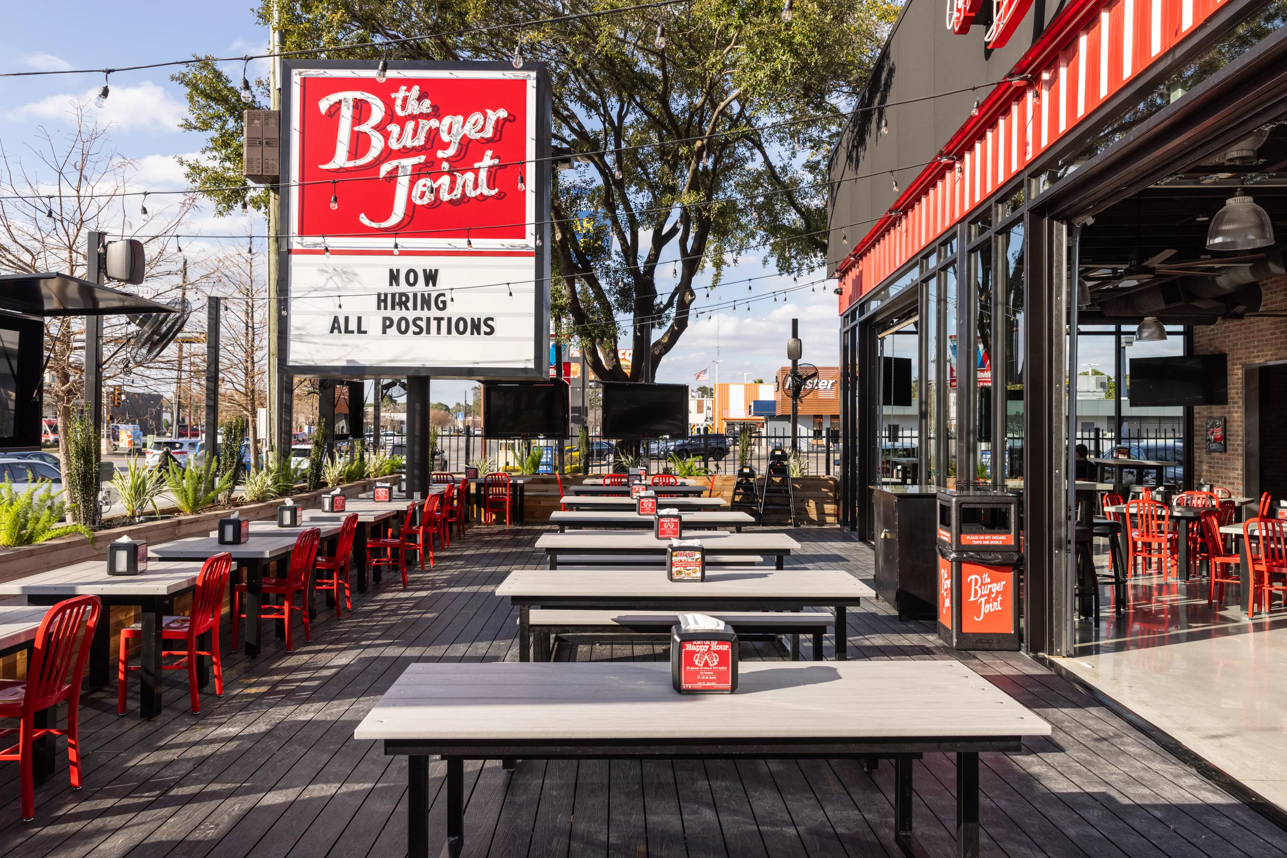 Outdoor seating area of a burger restaurant with long gray tables, red chairs, string lights, and a large sign that says 'The Burger Joint' with 'Now Hiring All Positions' underneath.