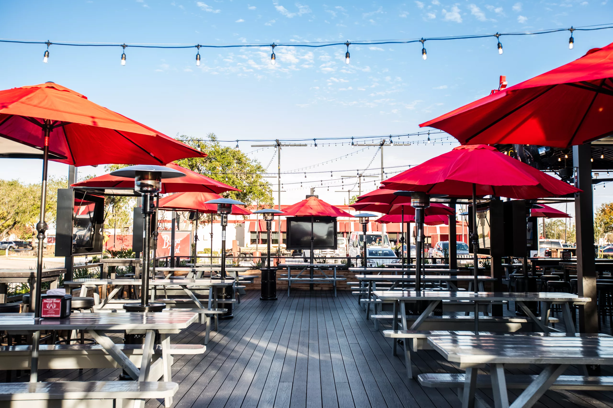 Outdoor patio with red umbrellas over picnic tables and string lights under a clear blue sky
