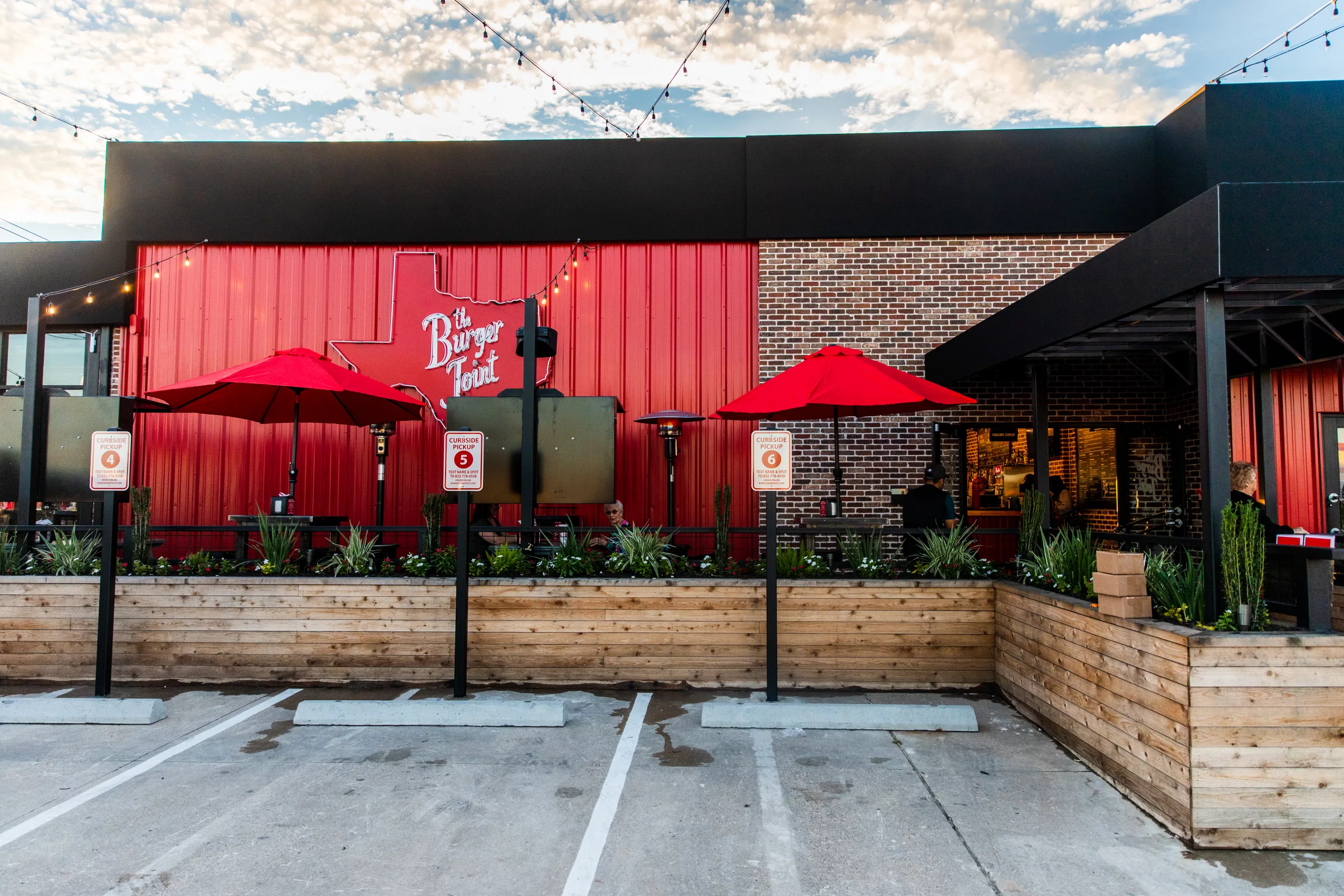 Exterior of a casual restaurant with a red wall, brick accent, and black awning; outdoor seating with red umbrellas and string lights.