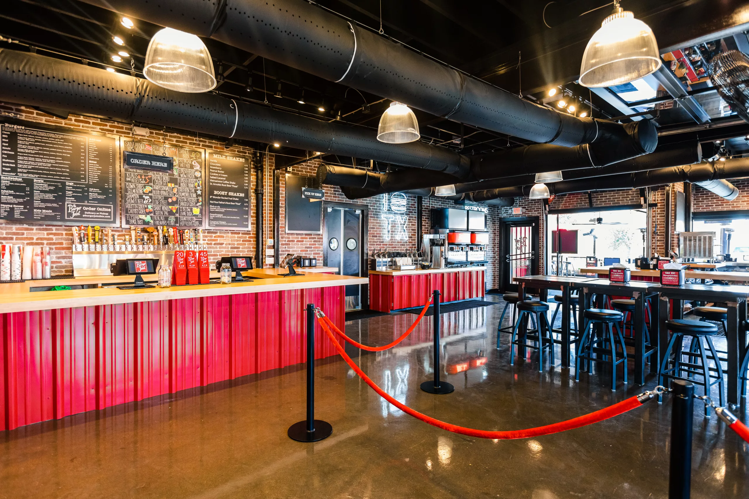 Interior of a casual burger restaurant: brick walls, black menu boards, red corrugated counter front, and red rope barriers at the front.