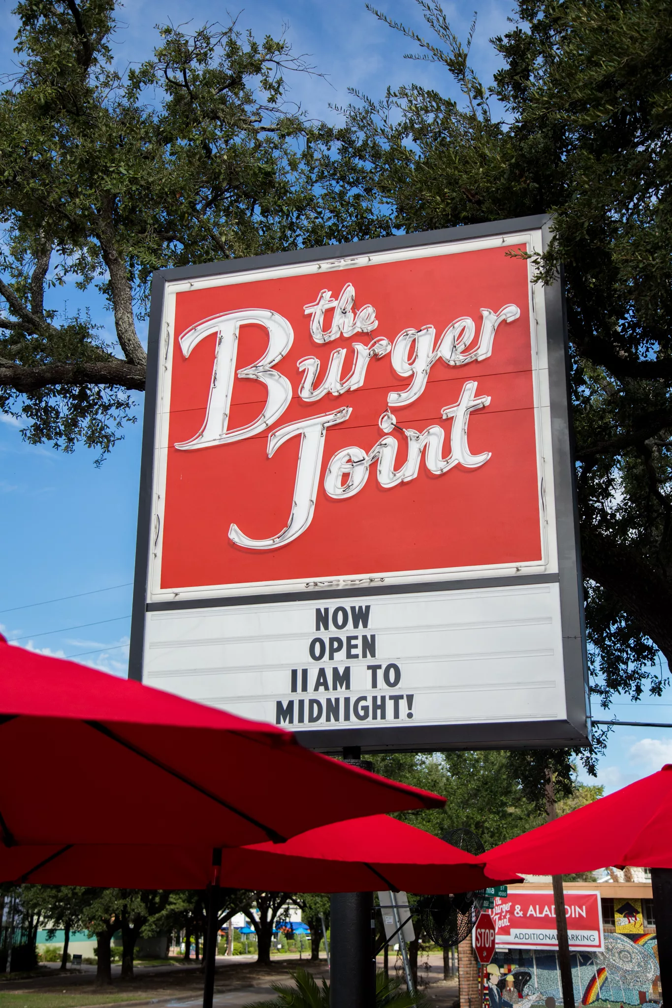 Red signage with white cursive 'The Burger Joint' frame, trees and blue sky behind.
