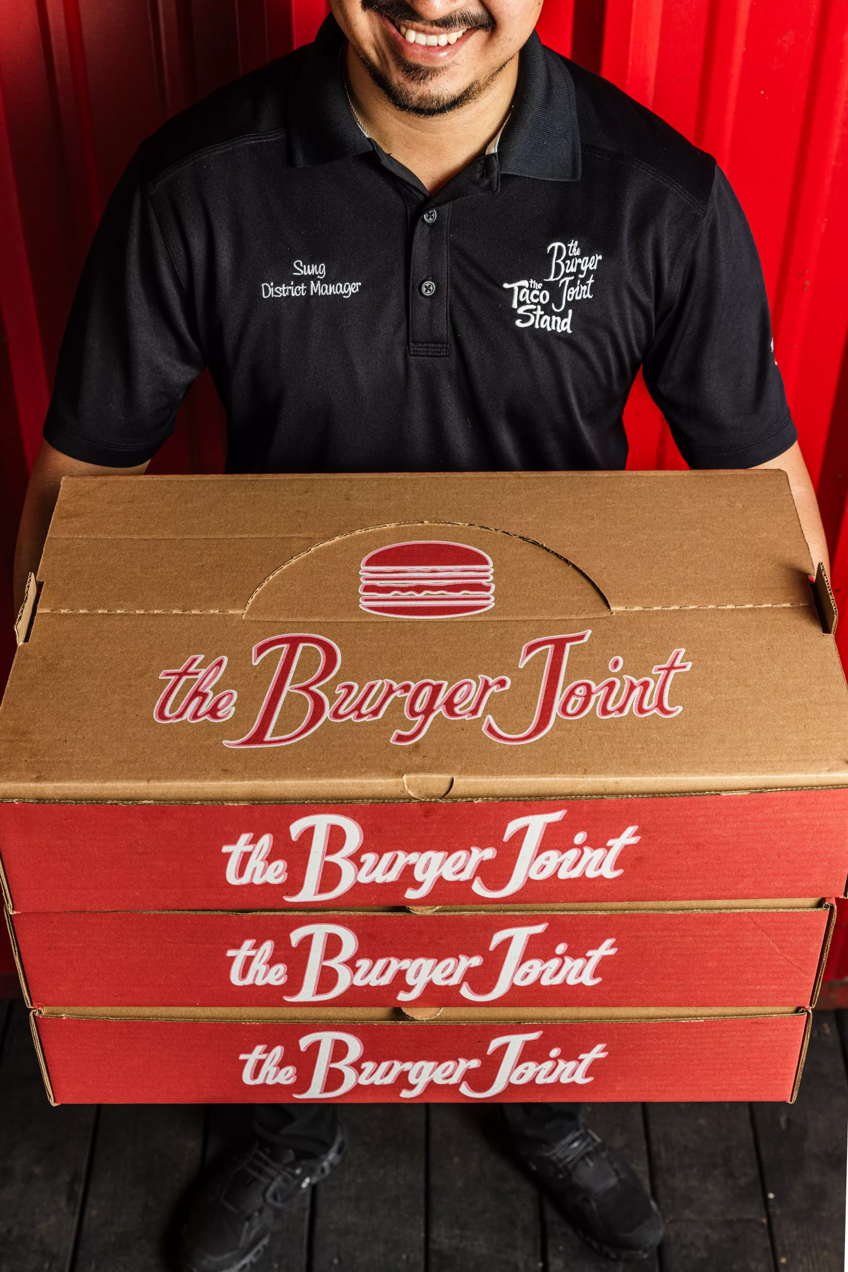 Man in a black polo with embroidered 'Sung District Manager' standing behind stacked The Burger Joint boxes against a red backdrop.